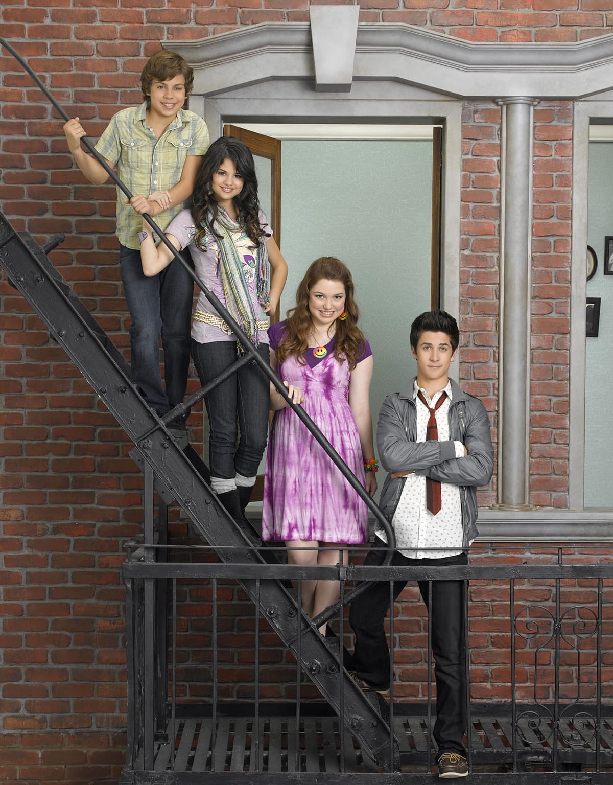 Jake T. Austin, Selena Gomez, Jennifer Stone, and David Henrie pose on a fire escape for Wizards of Waverly Place, with Gomez adding flair to her jeans-and-tee combo with layered scarves and boots, while Henrie keeps it sharp in loafers and a loosely knotted tie