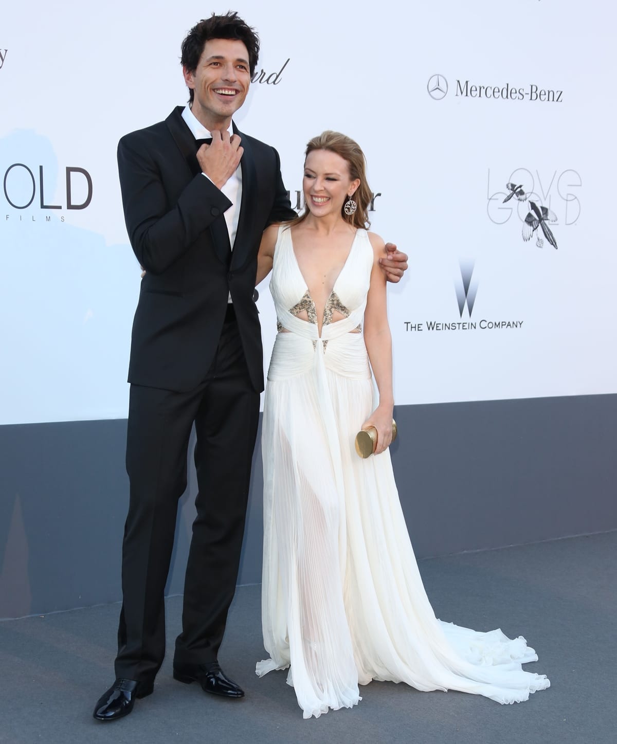 Kylie Minogue and Andrés Velencoso attend a red-carpet event together, with Kylie glowing in a flowing white gown featuring a plunging neckline and pleated skirt beside her sharply dressed partner in a classic black tuxedo