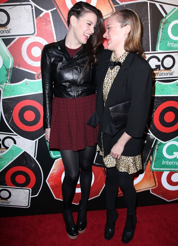 Liv Tyler towers over Chloë Sevigny in sky-high platform clog boots, using the exaggerated height and chunky sole to toughen a burgundy pleated dress and sleek leather moto jacket—an effortless study in feminine-meets-biker contrast