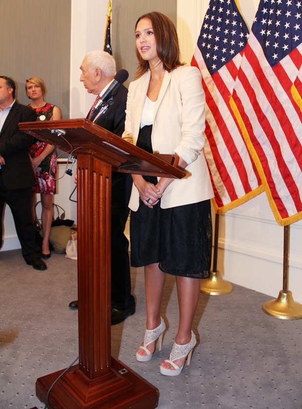 Jessica Alba speaks at a press conference, wearing a sand-colored Ann Taylor jacket and a black Jane Oh lace skirt, complemented by intricate white lace Elie Saab platform sandals
