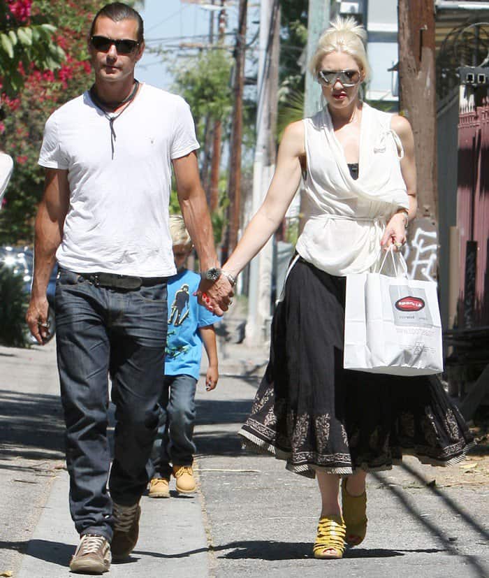 Gwen Stefani rocks a white wrap top with an ankle-length black embroidered skirt, paired with bold yellow L.A.M.B. Camden bootie sandals, while Gavin Rossdale complements her style in a casual yet sleek ensemble during a family outing