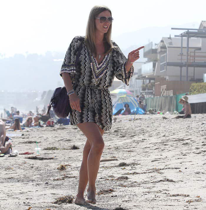 Nicky Hilton poses barefoot on a sandy beach, wearing a monochrome zigzag-patterned tunic dress with beaded details, large sunglasses, and a dark shoulder bag