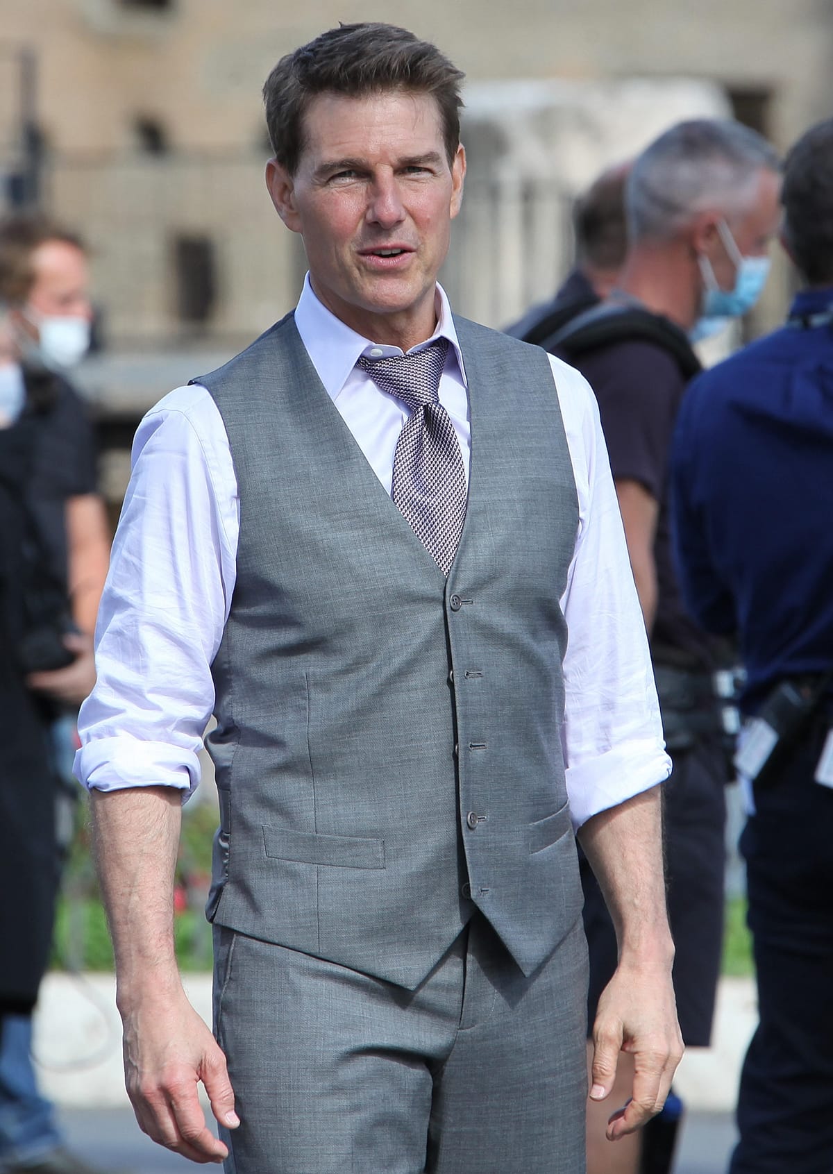 Tom Cruise looks effortlessly sharp on set in a tailored gray three-piece suit with a vest, styled over a crisp white shirt with rolled sleeves and a textured tie—his polished yet relaxed outfit balances Hollywood sophistication with behind-the-scenes ease