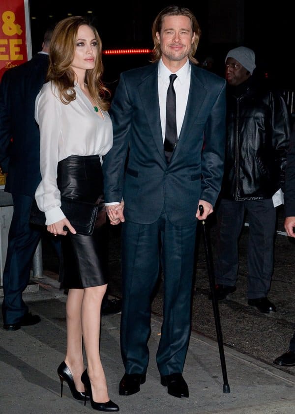 Angelina Jolie exudes elegance in a white silk blouse, black leather pencil skirt, and black pumps, while Brad Pitt complements her look in a sharp navy suit, white dress shirt, black tie, and formal dress shoes, accessorized with a cane