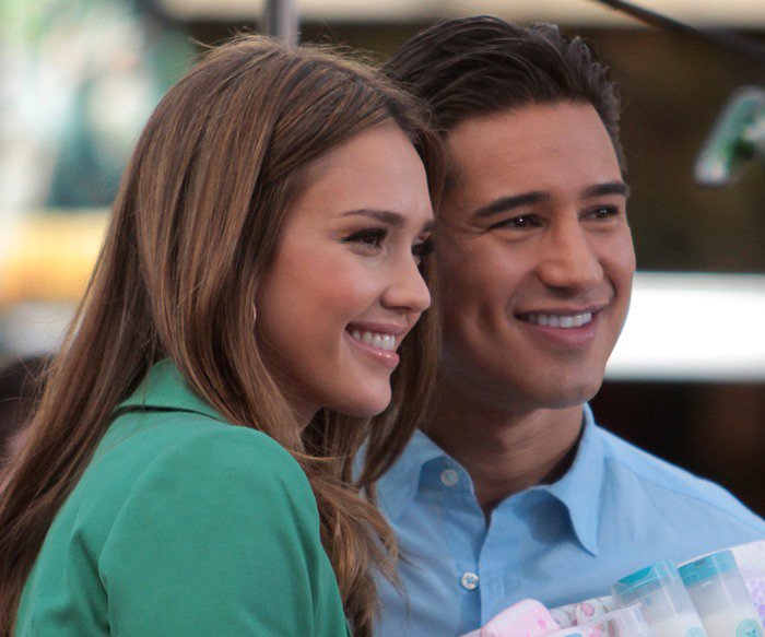 Jessica Alba poses beside Mario Lopez in a polished daytime look anchored by a mint-green blazer and flared dark-wash jeans
