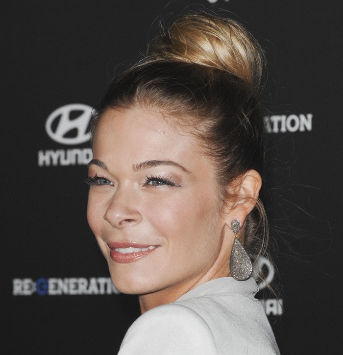 LeAnn Rimes wears her hair in a sleek high bun, accentuating her glowing skin, subtle shimmery eye makeup, and glossy nude lips, while accessorizing with glittering drop earrings
