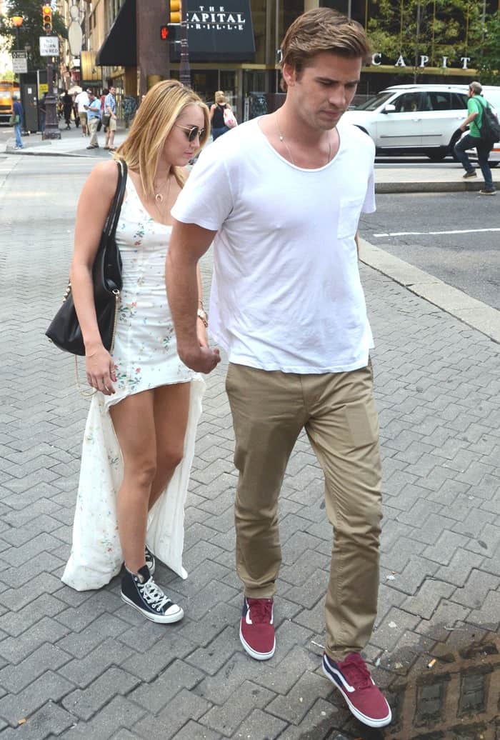 Miley Cyrus donning a chic Kate Moss for Topshop asymmetric dress, complemented by a Valentino studded leather tote, Michael Kors rose gold chronograph watch, Ray-Ban round sunglasses, and classic black Converse All Star Core Hi sneakers