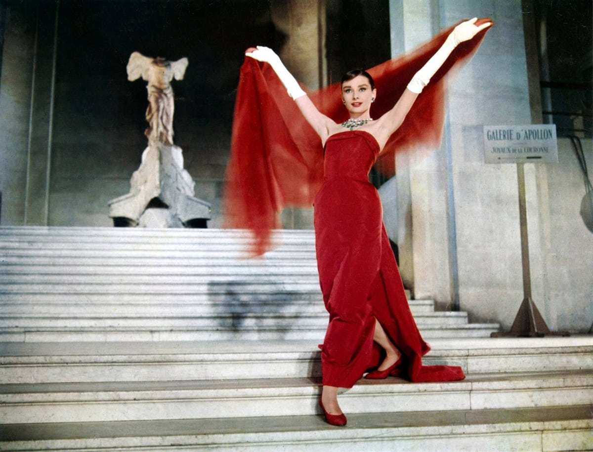 In the 1957 movie Funny Face, Audrey Hepburn, as bookstore clerk turned fashion model Jo Stockton, dazzles in a flowing red Givenchy gown, matching red heels, and a billowing red scarf while posing on the grand staircase of the Louvre