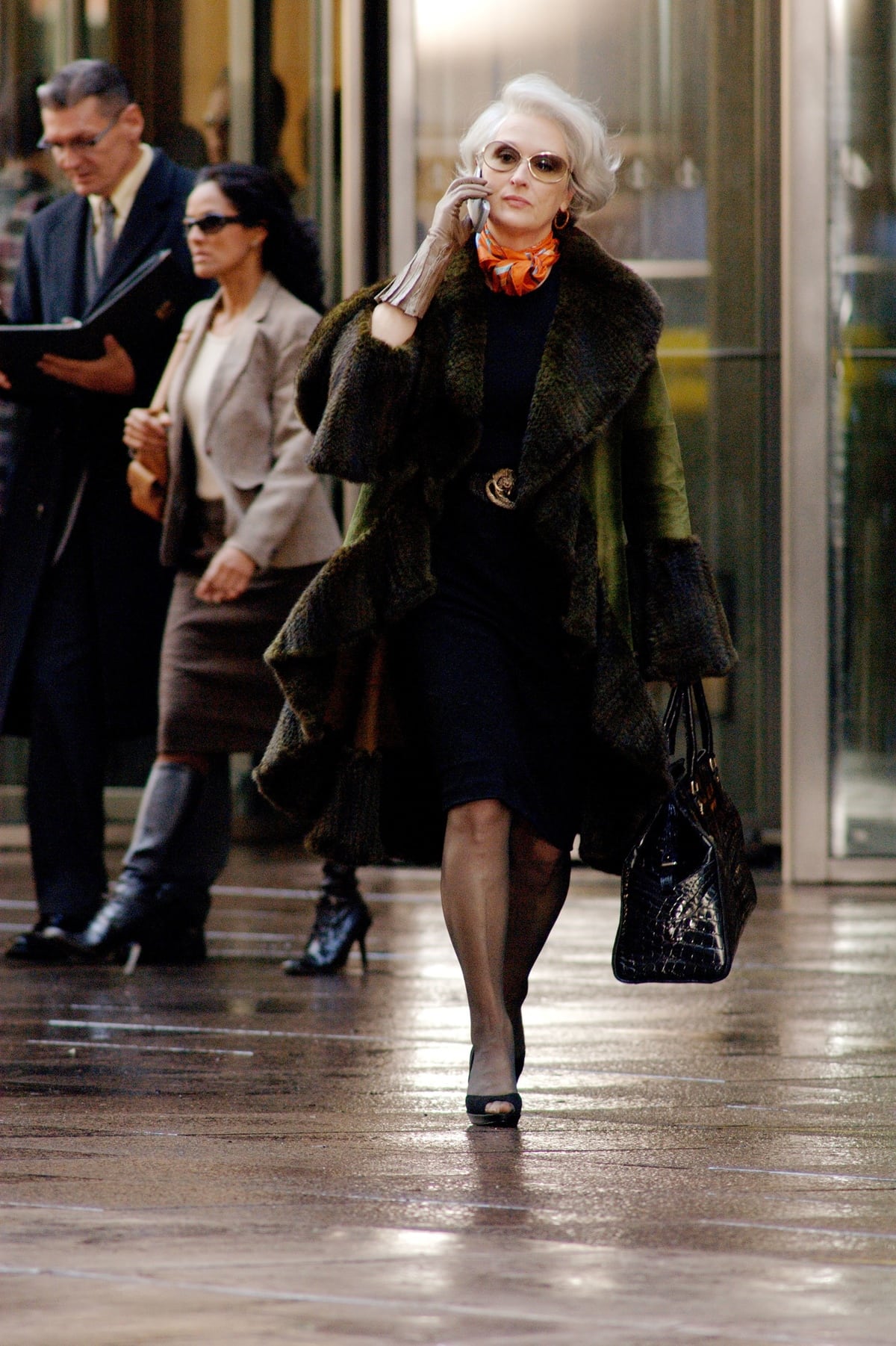 Meryl Streep, as Miranda Priestly in The Devil Wears Prada, commands the sidewalk in a luxurious green fur-trimmed coat, an orange Hermès scarf, a sleek black sheath dress, and classic black peep-toe heels