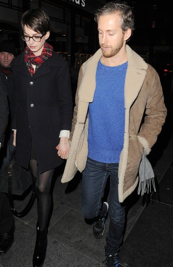 Anne Hathaway embraces polished cold-weather style in a dark tailored coat, plaid tartan scarf, opaque tights, and sleek black ankle boots while walking hand in hand with Adam Shulman in London