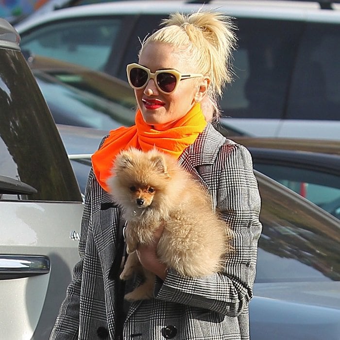 Cradling her fluffy pup, she sharpens a classic checked coat with cat-eye sunglasses and a vivid neon scarf, turning a casual school run into a confident, high-contrast style moment