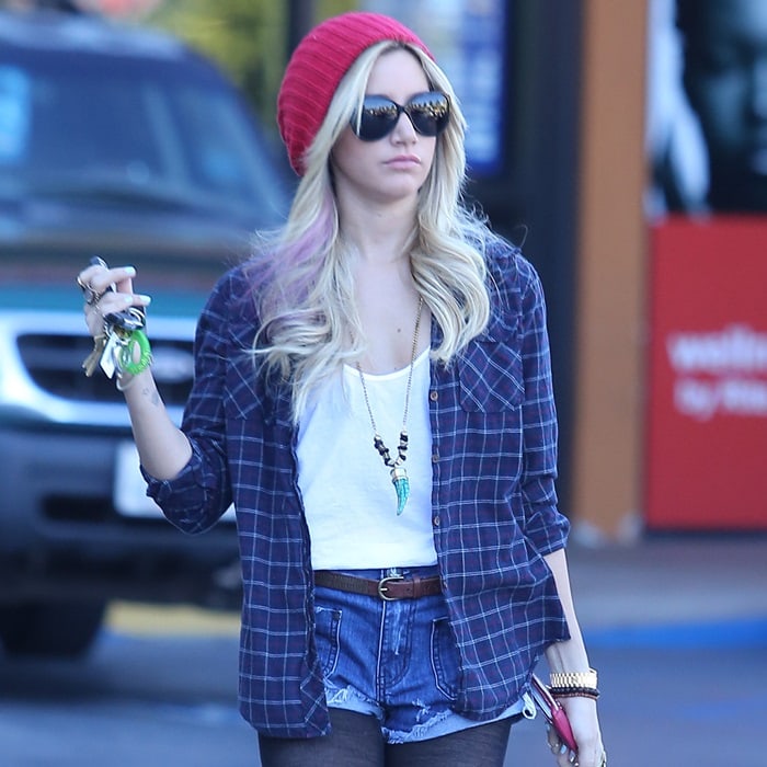 Ashley Tisdale is seen wearing a plaid shirt over a white tank top, jean shorts, and a red beanie