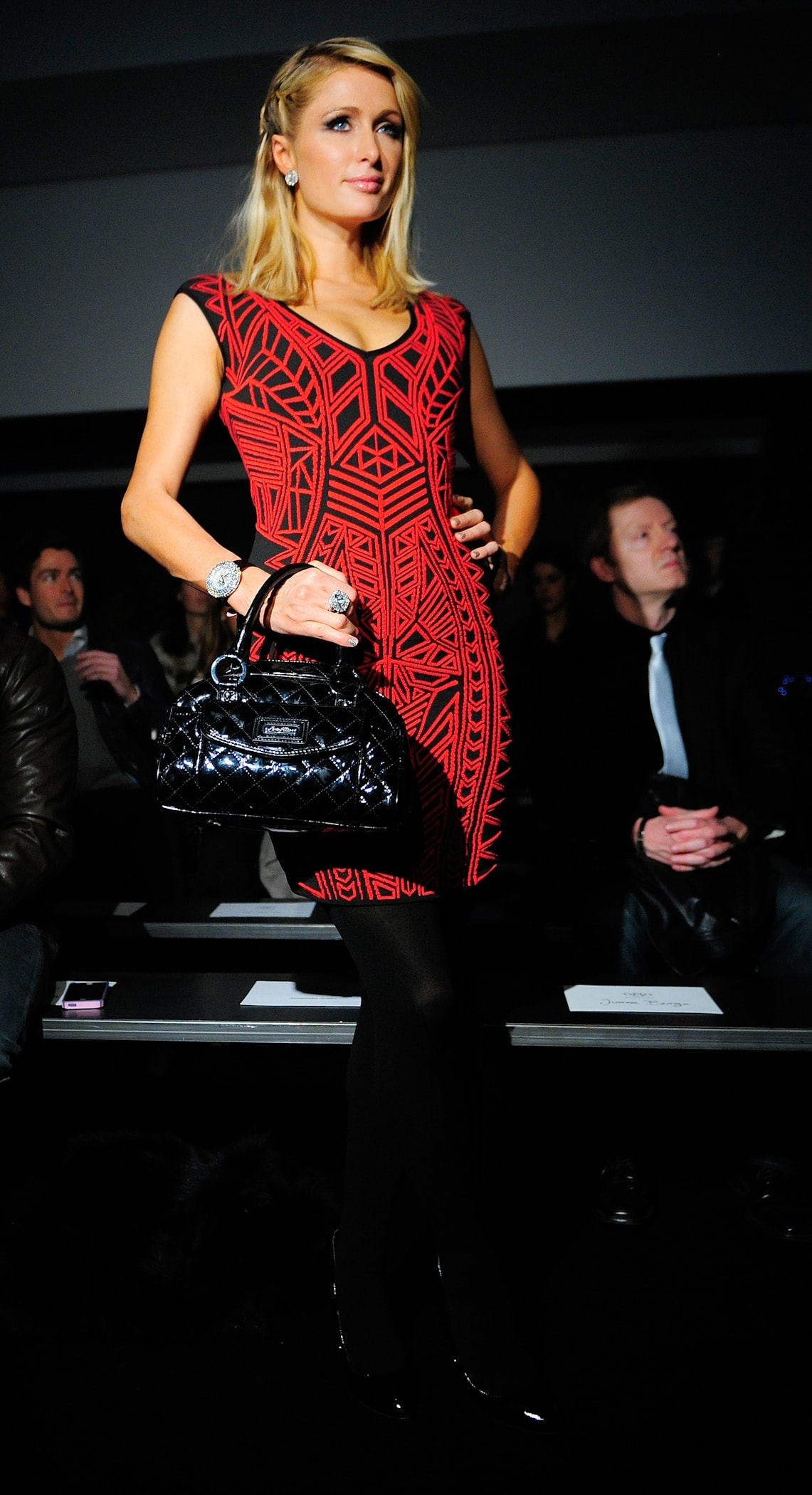 Paris Hilton stuns in a bold red and black geometric dress, paired with sleek black tights and a glossy quilted handbag for a striking front-row fashion moment