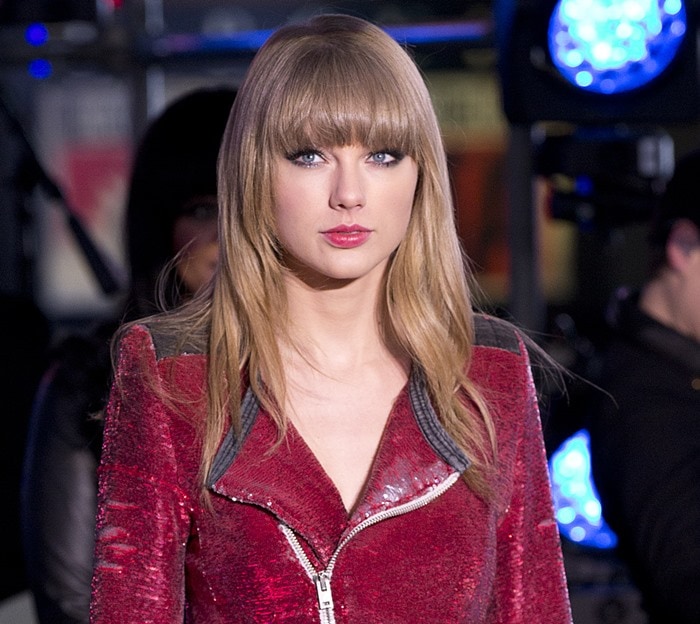 Taylor Swift wears her signature long blonde hair with full, blunt bangs and soft layers, complementing her bold red moto jacket