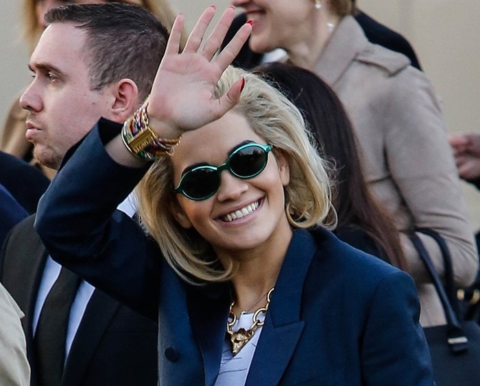 In her navy suit and signature accessories, Rita Ora waves to the crowd of fans and paparazzi outside the Burberry fashion show