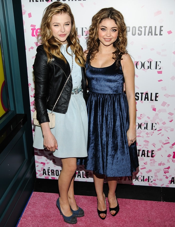 Sarah Hyland sparkles in a navy velvet alice + olivia by Stacey Bendet dress, the softly cinched waist and rich texture elevating the youthful silhouette, finished with classic black peep-toe heels that add a subtle lift and polished contrast as she poses alongside Chloë Grace Moretz