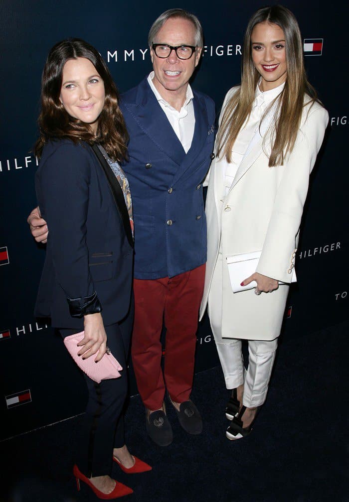 Drew Barrymore, Tommy Hilfiger, and Jessica Alba pose together at the brand’s West Coast flagship opening, showcasing coordinated tailoring—Barrymore in sharp navy with red pumps, Hilfiger in classic Americana layers, and Alba in a crisp all-white suit finished with graphic black-and-ivory heels