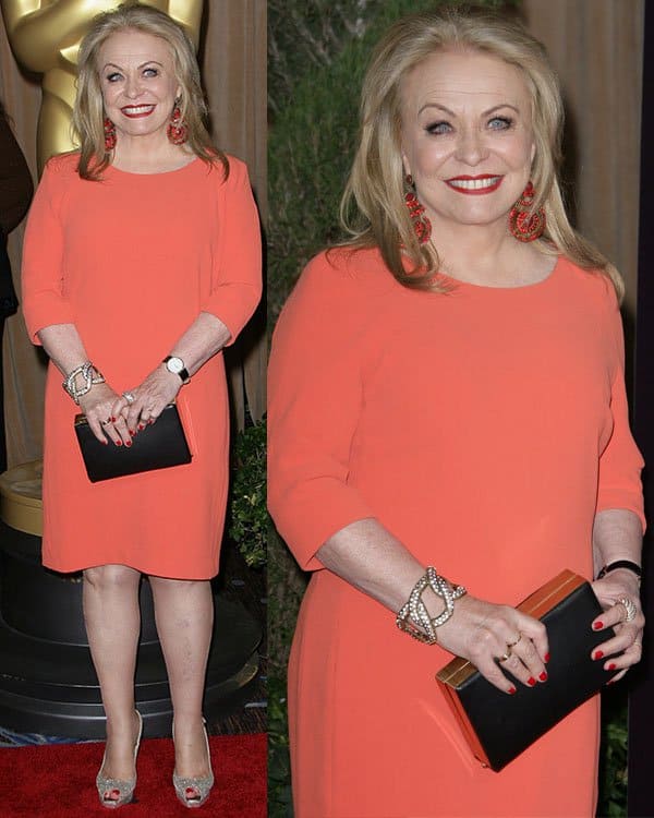 Jacki Weaver wears a bold orange knee-length dress with three-quarter sleeves, accessorized with red statement earrings, a black clutch, silver bracelets, and metallic peep-toe heels