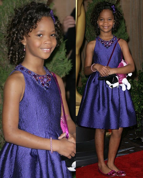 Quvenzhané Wallis wears a sleeveless purple satin dress with a jeweled neckline, accessorized with a matching headband, a playful puppy purse, and glittery pink flats