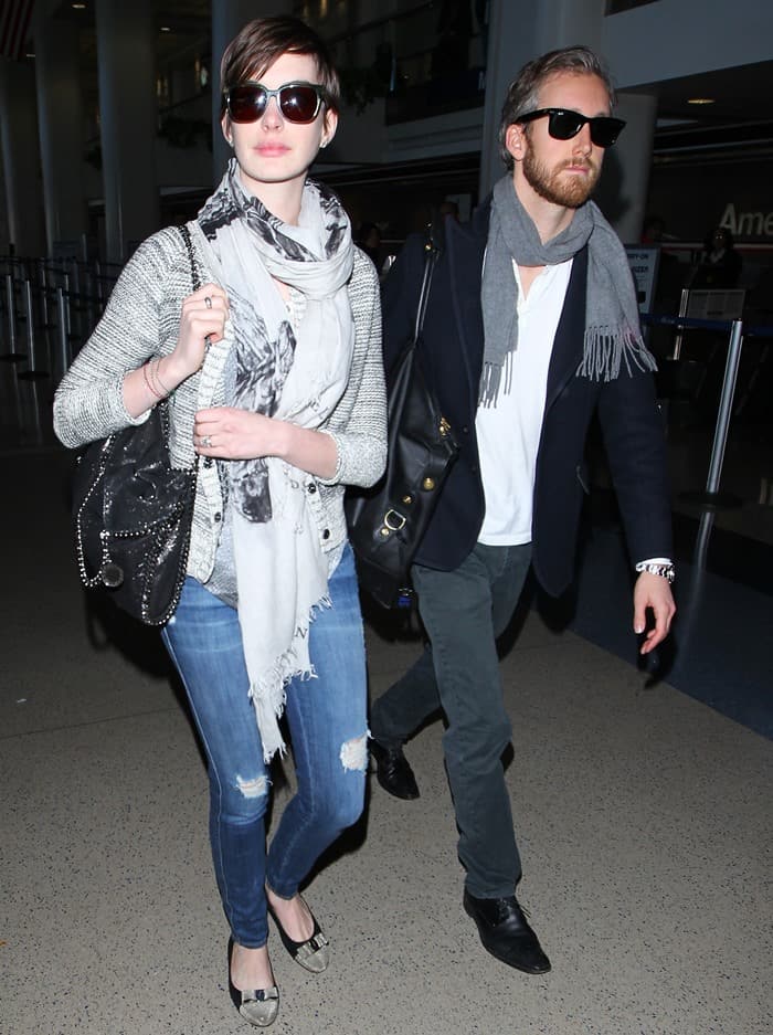 Anne Hathaway and Adam Shulman coordinate understated airport style at LAX, pairing layered scarves and dark sunglasses with relaxed denim, loafers, and polished black leather shoes for an effortlessly cohesive travel look