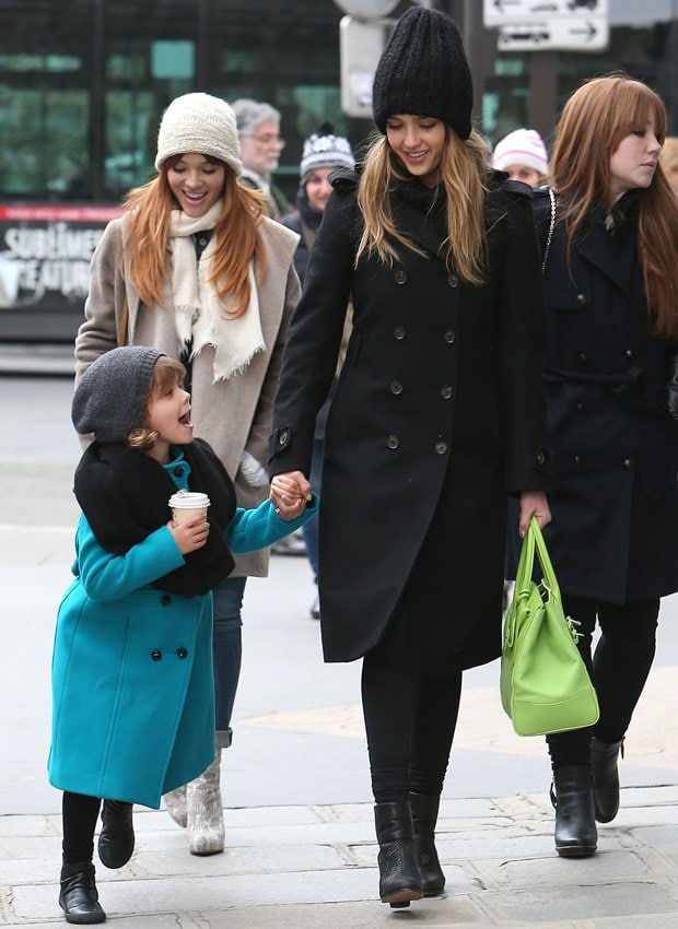 Smiling mid-stroll, Alba pairs her winter-black ensemble and ankle boots with a pop of lime-green handbag as Honor walks beside her in a teal coat, knit hat, and practical boots suited for city exploring