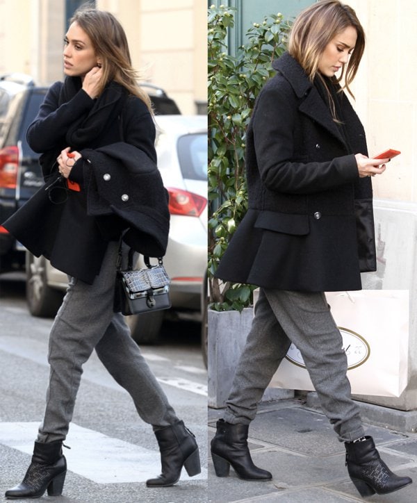Jessica Alba keeps her Paris shopping look understated in a black coat and gray jogger pants, elevating the ensemble with standout accessories — a textured satchel bag with a geometric flap, sleek Rag Bone “Newbury” booties, and a bold red phone case adding a subtle pop of color
