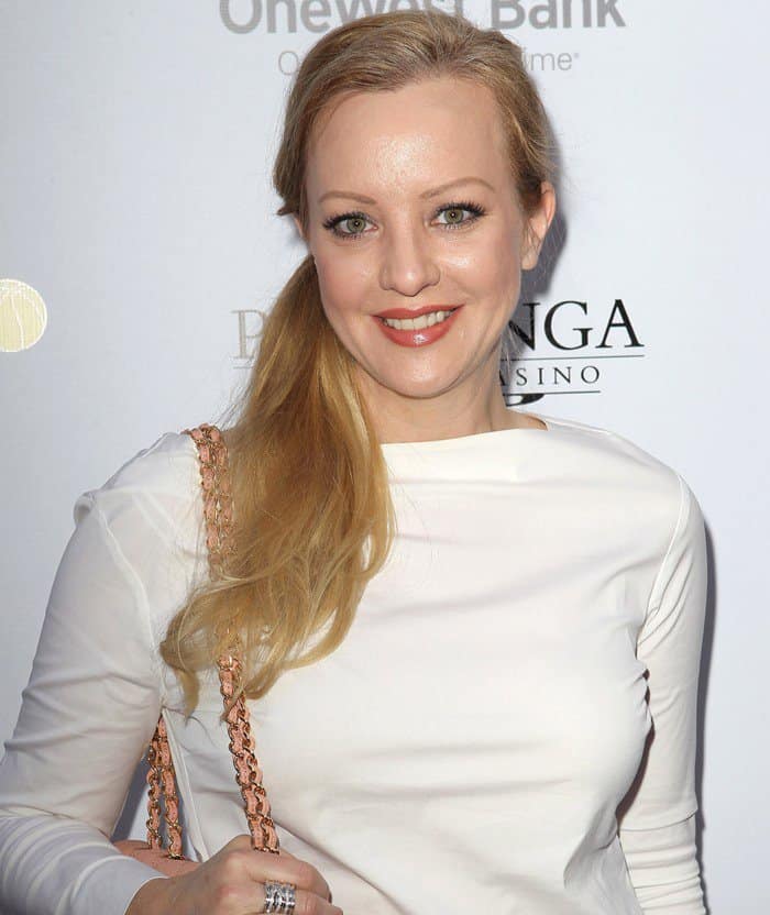 Wendi McLendon-Covey smiles for a picture while wearing a white long-sleeve top and carrying a pink quilted handbag with a chain strap