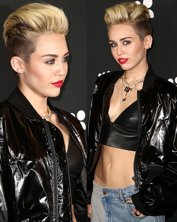 Miley Cyrus's bold look features a voluminous blonde mohawk and striking red lipstick, complemented by multiple necklaces, earrings, and rings
