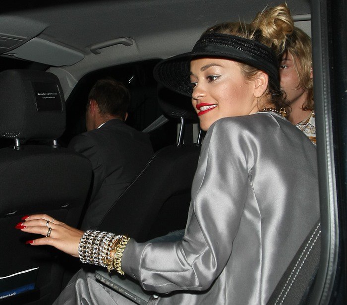 Rita Ora exudes edgy glamour in a silver Armani suit, layered gold jewelry, and a black straw visor, adding a sporty-chic twist to her polished studio look