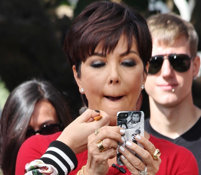 Kris Jenner in a red outfit holding her phone while having her makeup touched up during an Extra appearance