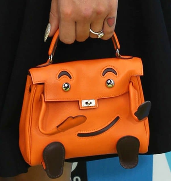 Detailing her playful side, Kelly Osbourne complements her sophisticated black dress with a charming vintage orange Hermès handbag featuring a smiley face, pictured here alongside her engagement ring from then-fiancé Matthew Mosshart