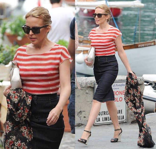 Kylie Minogue exudes effortless summer elegance in a red striped top tucked into a high-waisted denim pencil skirt, accessorized with chic cat-eye sunglasses, a crisp white clutch, and floral scarf for a touch of contrast, as she strolls through the picturesque streets of Portofino