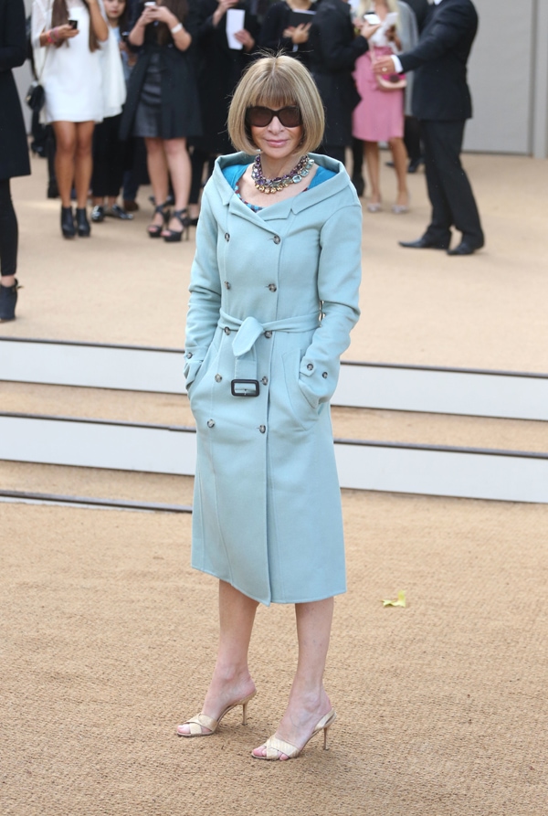 Anna Wintour radiates polished elegance in a belted pale blue trench coat paired with woven nude heeled mules and her signature oversized sunglasses