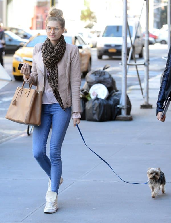 Jessica Hart walks her dog in New York City, effortlessly combining comfort and style with a beige moto jacket, blue skinny jeans, white high-top Converse sneakers, a chic leopard print scarf, and a tan Prada Saffiano Lux Tote handbag