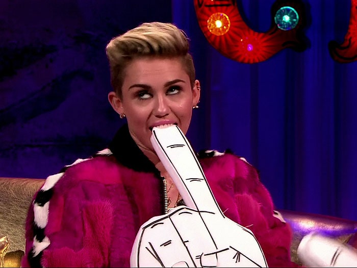 Miley Cyrus playfully bites a foam finger during her Alan Carr: Chatty Man appearance, offsetting the bold, textured pink jacket with a slick, patent bralette for contrast