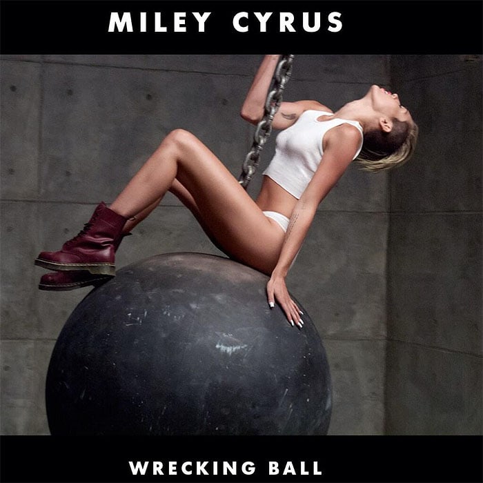 Miley Cyrus is pictured in a white crop top and underwear, paired with cherry-red Dr. Martens boots, perched on a wrecking ball for her "Wrecking Ball" single cover