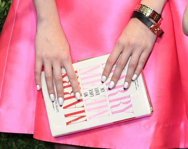 A stylish 'Marry Me' book clutch, perfectly paired with white-manicured nails and a vibrant pink dress, accessorized with a chic gold and black bracelet