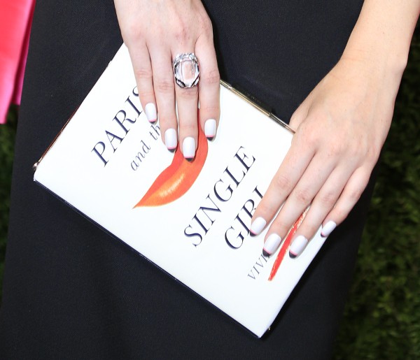 A close-up of the 'Paris and the Single Girl' book clutch, complemented by white-manicured nails and a statement ring, adding sophistication to a classic black outfit