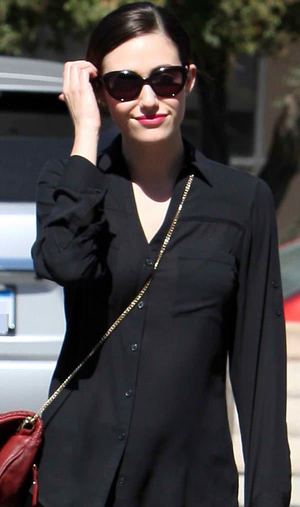 Emmy Rossum looks effortlessly chic in a black button-down top and cat-eye sunglasses, accessorized with a red Zadig Voltaire pochette and coral-red lips