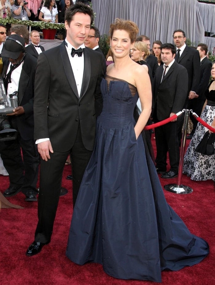 Sandra Bullock wore a floor-length navy Angel Sanchez gown at the 78th Academy Awards, likely paired with heels hidden beneath the full skirt, while Keanu Reeves complemented her look in a timeless black tuxedo