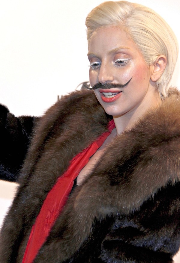 Lady Gaga poses with a dramatic Salvador Dali mustache and fur coat, showcasing her signature bold style at the 'ARTPOP' listening party