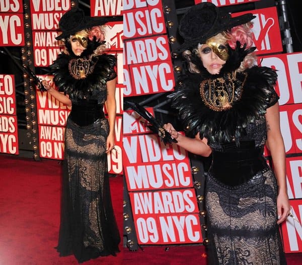 Lady Gaga arrives at the 2009 MTV Video Music Awards in New York City wearing a sheer black Jean Paul Gaultier lace gown layered over nude fabric and styled with an elaborate feathered collar, gold face mask, miniature top hat, and dramatic black accessories, turning the red carpet into a piece of theatrical fashion performance rather than traditional celebrity glamour on September 13, 2009