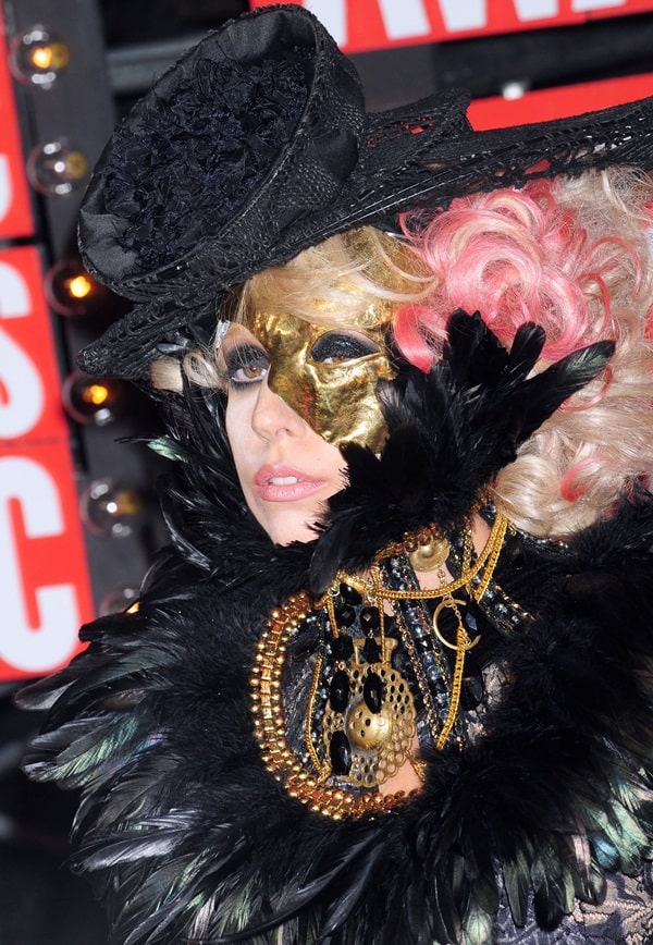 A close-up of Lady Gaga’s 2009 MTV Video Music Awards ensemble reveals the intricate theatrical styling behind the look, including a metallic gold half-mask, dramatic black feather collar, layered antique-style jewelry, lace textures, and a miniature black hat set against platinum curls streaked with pink