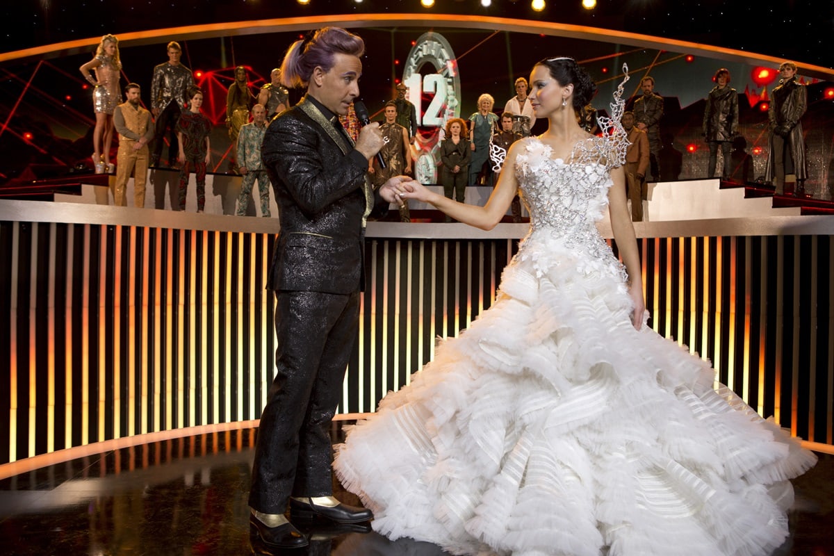 Jennifer Lawrence wears the iconic Tex Saverio “Mockingjay” wedding dress in The Hunger Games: Catching Fire, a dramatic white gown with a sculpted bodice and cascading ruffled skirt designed for the Capitol ceremony scene