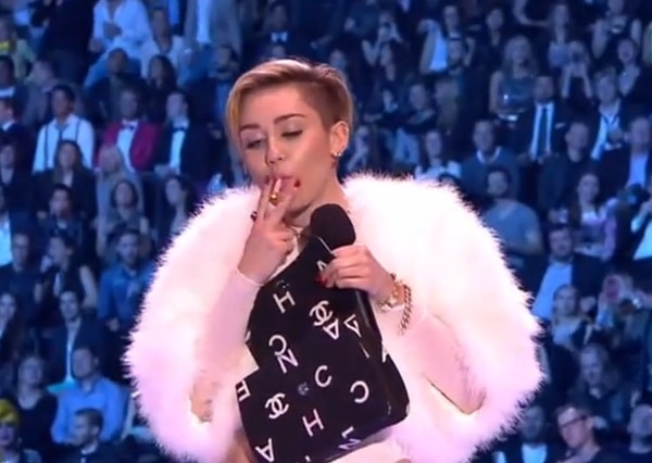 Miley Cyrus smoking a joint on stage at the MTV Europe Music Awards, wearing a white fur coat and holding a Chanel bag