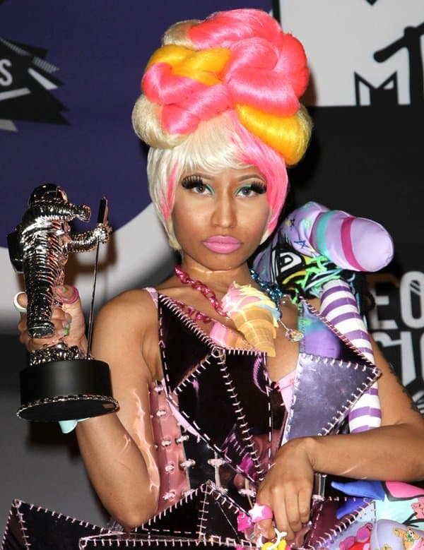 Nicki Minaj’s 2011 VMAs outfit combined sculptural fashion with toy-like fantasy styling, pairing a sharp-edged patent patchwork dress with mismatched printed tights, stuffed character accessories, and towering novelty platform shoes that reinforced her larger-than-life “Harajuku Barbie” aesthetic during the peak of her early pop crossover era
