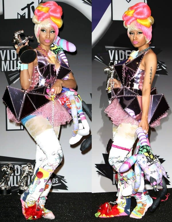 Nicki Minaj poses backstage at the 2011 MTV Video Music Awards holding her Moon Person trophy while wearing a chaotic candy-colored ensemble designed by Furne One, complete with a towering pink-and-yellow beehive wig, patchwork geometric dress, cartoon-inspired leggings, plush accessories, and exaggerated platform footwear that blurred the line between fashion, costume design, and pop-art performance in Los Angeles on August 28, 2011