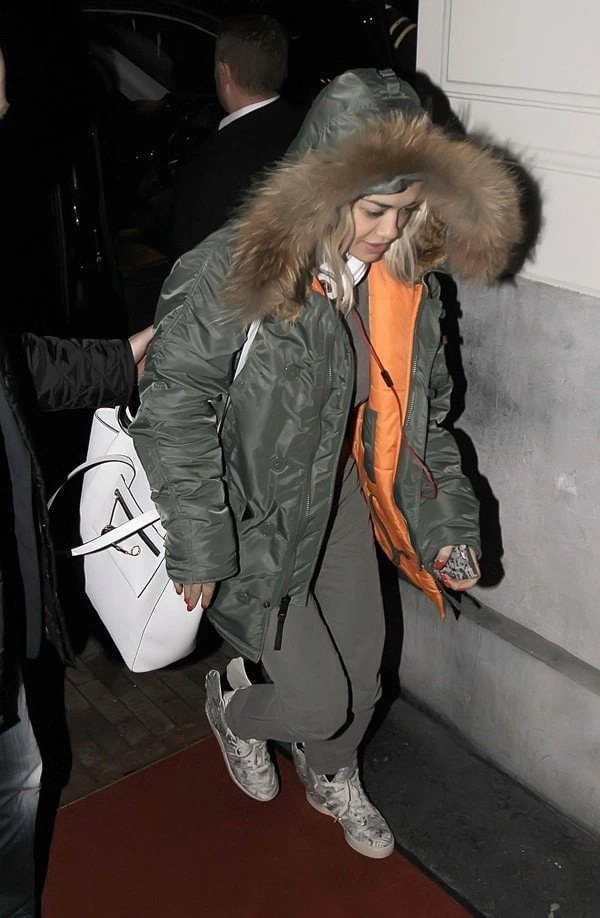 Rita Ora bundled up in a dark green parka with fur trim, styling it with relaxed-fit overalls, winged Jeremy Scott x Adidas sneakers, a white handbag, and matching Beats by Dre headphones for a playful, cozy look