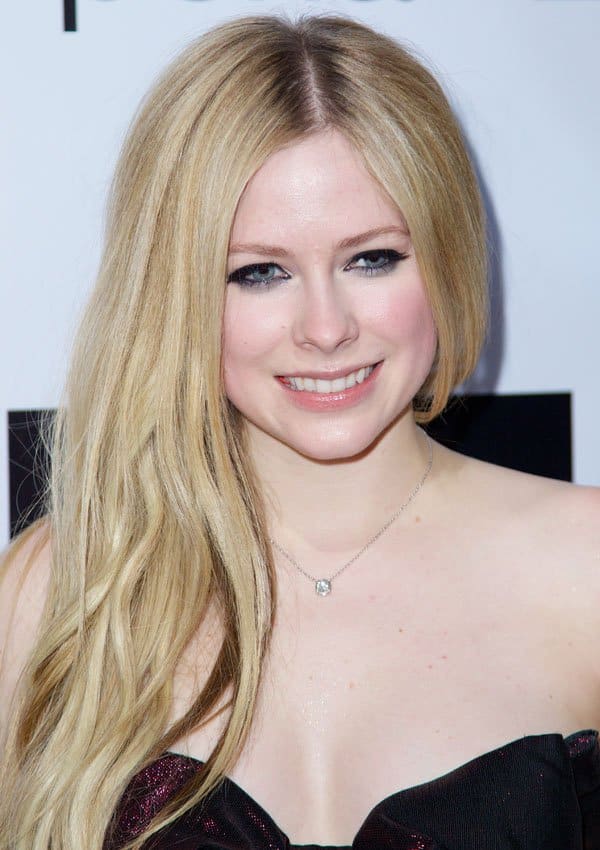 Avril Lavigne smiles brightly, highlighting her flawless teeth and glowing makeup with subtle pink blush and nude lips, complemented by her long, loose waves