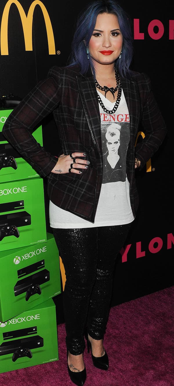 Demi Lovato beside a stack of unopened Xbox One boxes, looking stunning in sequined jeans and a plaid jacket